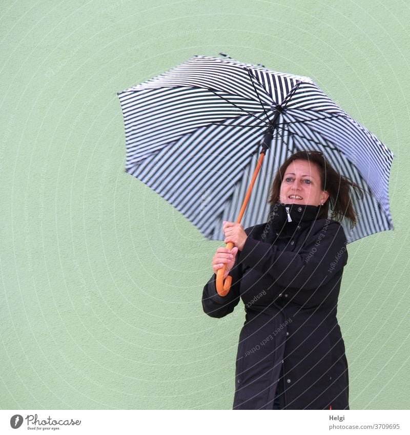 lächelnde Frau im schwarzen Mantel steht vor einer hellgrünen Wand und hält einen blau-weiß-gestreiften Schirm Mensch weiblich feminin Regenschirm langhaarig