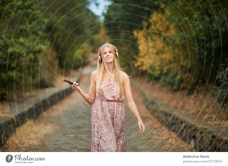 Lächelnde Frau mit Smartphone und Kopfhörer zu Fuß im Park Glück zuhören benutzend Musik Sommer genießen jung Browsen Gasse Weg Gerät Apparatur Lifestyle Stil