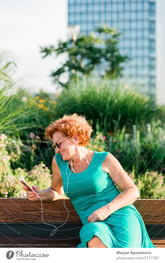Reife Frau, die im Park die Musik auf ihrem Smartphone hört 60s Erwachsener Lebensalter gealtert schön Bank lässig Kaukasier heiter Großstadt Konnektivität