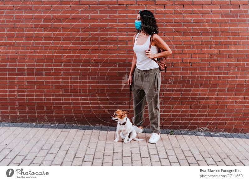 junge Frau, die mit Schutzmaske im Freien spazieren geht, daneben süßer Jack-Russell-Hund. Neues normales Konzept Straße neue Normale jack russell Haustier