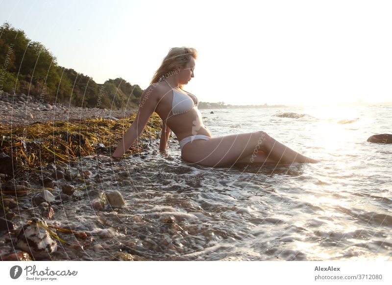 Portrait einer jungen, blonden Frau im Bikini, die in der Ostsee- Brandung sitzt junge Frau Mädchen 18-20 Jahre schlank schön lockig sinnlich natürlich