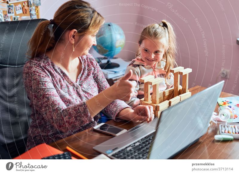 Eine Mutter, die während eines Video-Chat-Call-Stream-Online-Kurses von zu Hause aus auf einem Laptop arbeitet, während ihre Tochter mit einem Ziegelsteinspielzeug spielt. Frau sitzt am Schreibtisch vor dem Computer und schaut auf den Bildschirm