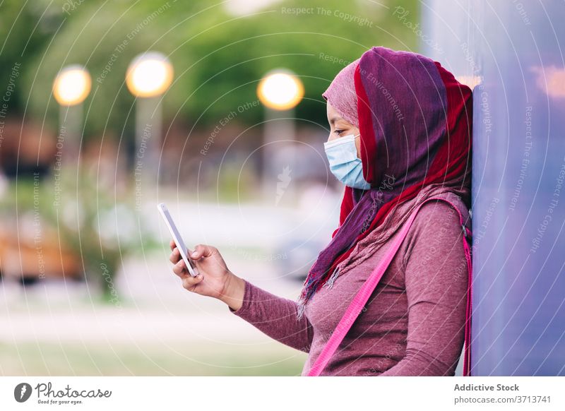 Sportliche Frau in Maske mit Smartphone auf der Straße Sportlerin Großstadt benutzend Hijab Mundschutz medizinisch Coronavirus Sportbekleidung arabisch ethnisch