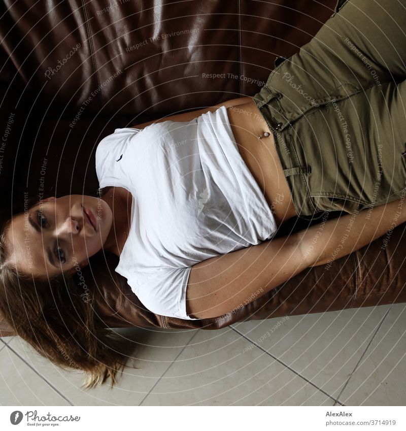 Portrait einer jungen Frau, die auf einer braunen Leder- Couch liegt und nach oben in die Kamera blickt sportlich feminin Empathie Gefühle emotional Porträt
