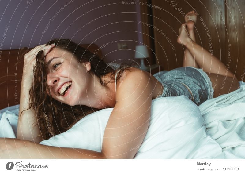 Eine junge Frau mit brünetten Haaren liegt auf dem Bauch im Bett. Das Mädchen lacht und hat einen Arm am Kopf. Ihre Füße sind in der Luft. Sie sieht glücklich aus.