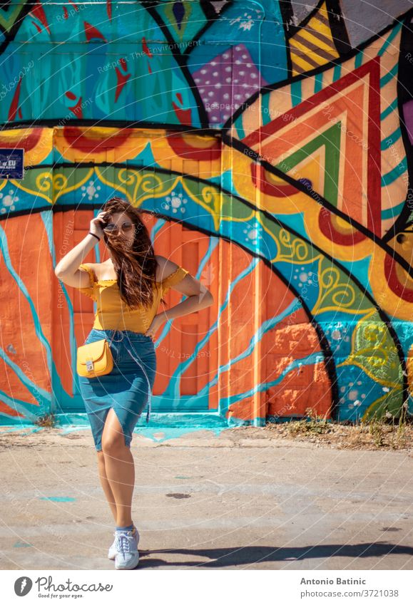 Attraktives brünettes Mädchen in einem blauen Rock und einem orangefarbenen Top posiert an einem Sommertag mit einer bunten Wand im Hintergrund. Gelbe Geldbörse, stilvolle Einflussnehmerin beim Reisen und Modellieren