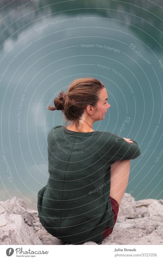 Frau sitzt am Ufer eines Petrolfarben Bergsee ufer Lünersee dunkelgrün braune Haare Zopf Sommer Sommerurlaub entschleunigung Ruhe Zeit Spiegelung im Wasser
