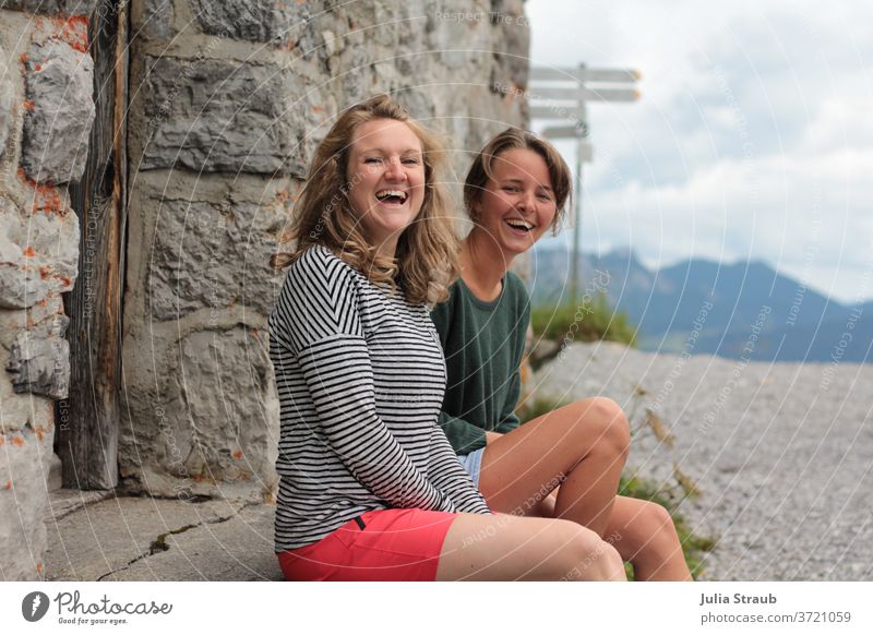 zwei junge Damen sitzen lachend auf der Stufe vor einem Steinhaus in den Bergen Berge u. Gebirge Wanderung wandern Pause Pause machen Spaß haben spass Freude