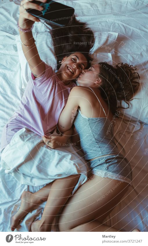 Portrait von zwei hübschen Freundinnen, die sich am Handy selbstständig machen, in einem Bett mit weißen Laken liegend, glücklich lächelnd. lesbisch Selfie
