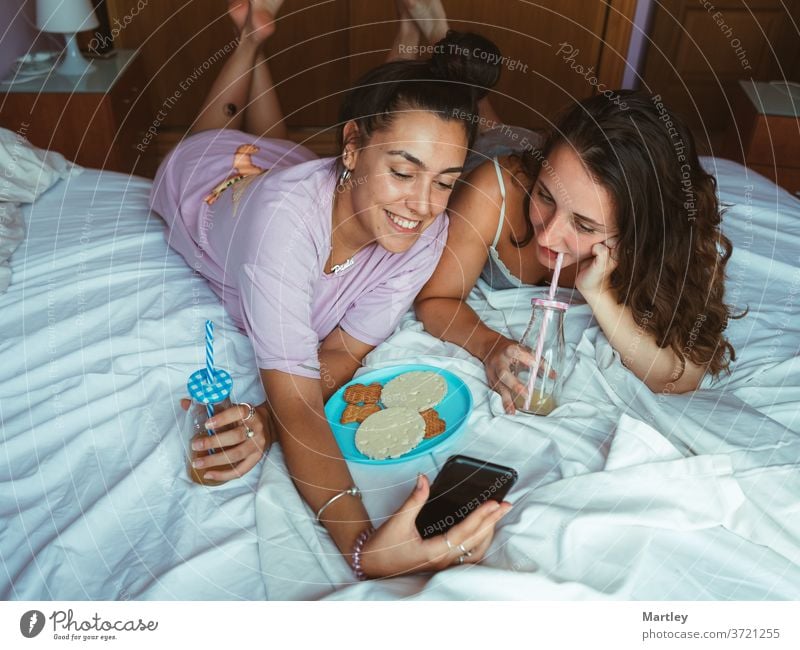 Zwei Freundinnen frühstücken im Bett eines Hotelzimmers, liegen zusammen und haben Spaß beim Reden und mit Smartphones beim Surfen im Internet. Frau Menschen