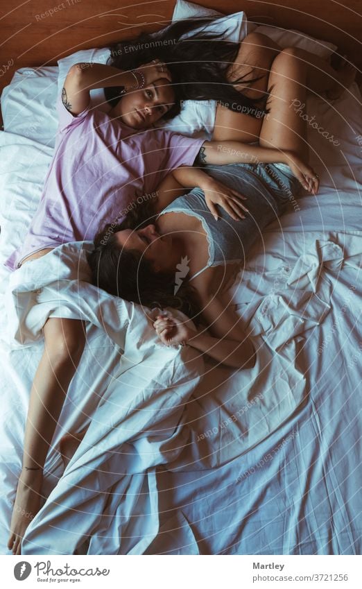 Zärtliche Szene von zwei jungen Frauen, die in einem Bett mit weißen Laken liegen und mit nachdenklichem Ausdruck kuscheln. lesbisch Selfie zu Hause