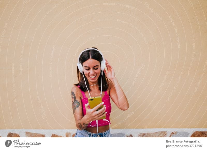 Fröhliche junge Frau im Freien, die mit Handy und Headset Musik hört. Technologie und Lebensstil Musik hören Glück Tattoo Technik & Technologie Großstadt urban