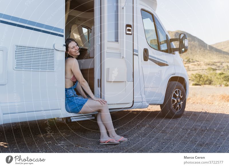 Lächelnde junge Frau, die an einem sonnigen Tag auf der Tür ihres Wohnmobils sitzt, das in einer wüstenartigen Naturlandschaft geparkt ist Lebensstile