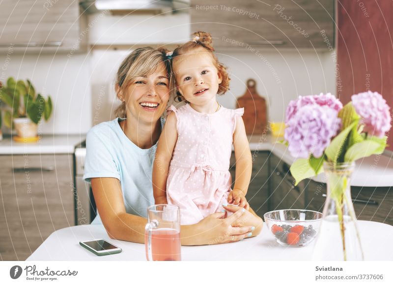 Porträt einer attraktiven jungen Frau und ihrer kleinen Tochter, die in der Küche sitzen und lächeln. Auf dem Tisch steht ein Bouquet aus Hortensien, Beeren und einem Glas Saft, Seitenansicht