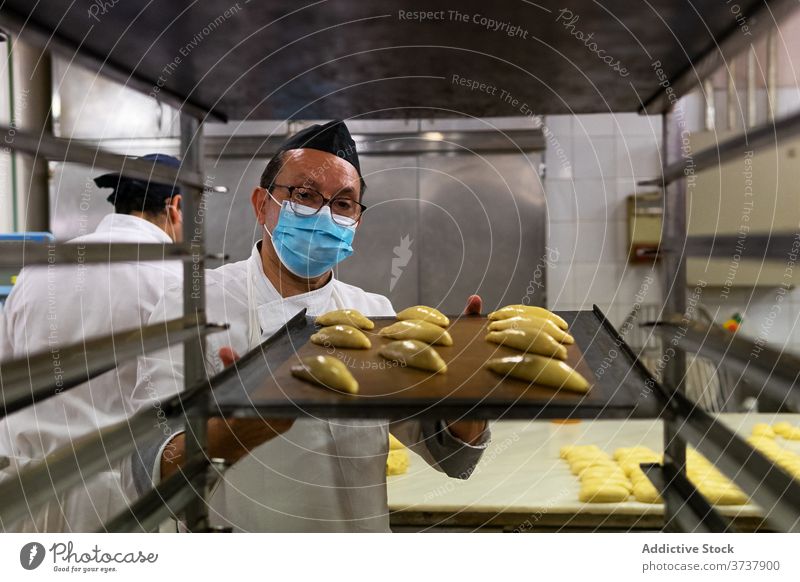 Ethnischer Mann beim Brötchenbacken in der Bäckerei Gebäck Koch vorbereiten medizinisch Mundschutz Coronavirus Teigwaren roh männlich ethnisch geschmackvoll