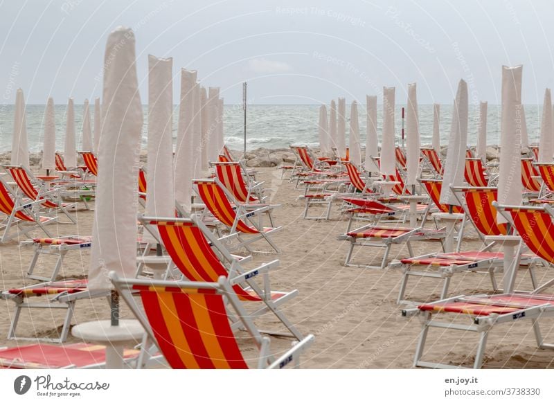 kein Sommerfeeling mehr - zugeklappte Liegestühle, Sonnenschirme und Klappstühle am Sandstrand in Italien vor einem Meer mit Wellen bis zum Horizont und bedecktem Himmel