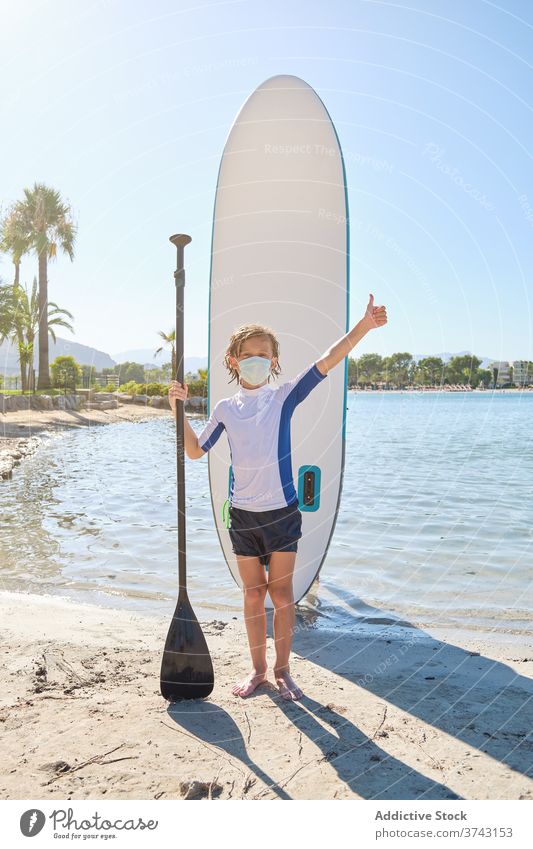 Kind mit einer Maske vor einem Paddel Surfbrett stehen, während eine Geste zu tun, um fein mit der Hand sein Donnern Atem Pandemie Prävention anhaben Seuche