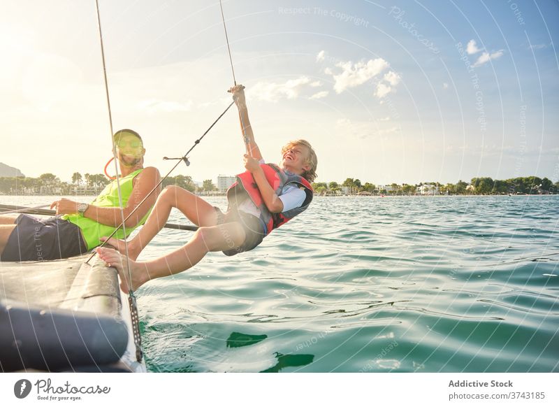 Junge mit Schwimmweste lachend und ein Seil von einem Boot ergreifend, um Gegengewichte zu machen, die auf das Meer hinaushängen Lachen gestikulieren