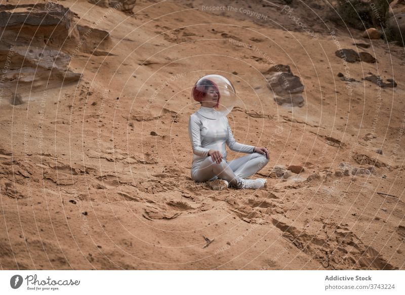 Lächelnde futuristische Frau im Splitteranzug Raum Astronaut Anzug positiv jung sitzen Felsen Stein Kosmonaut Tracht Konzept Silber Schutzhelm ruhen
