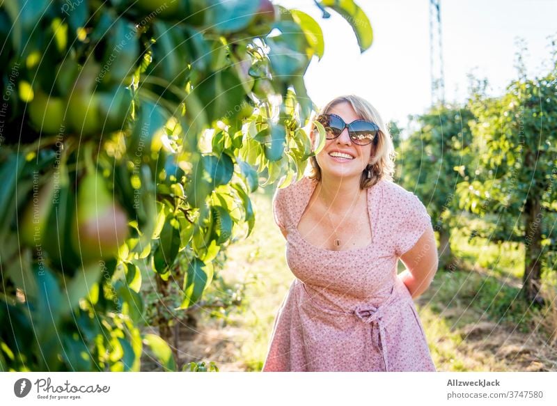 junge blonde Frau mit Sonnenbrille und rosa Kleid lacht in die Kamera in einer Obstplantage Junge Frau Sonnenuntergang sonnebrille Leichtigkeit Achtsamkeit