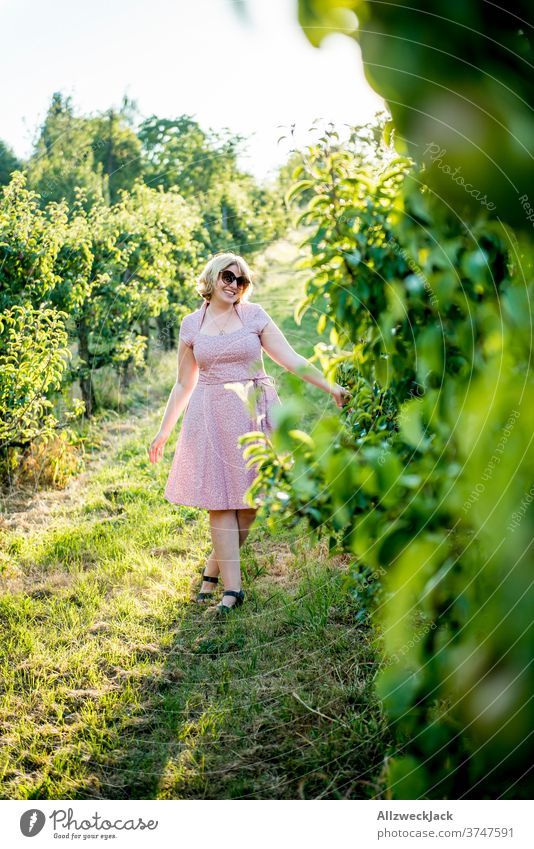 junge blonde Frau mit Sonnenbrille und rosa Kleid läuft fröhlich durch eine Obstplantage Junge Frau Sonnenuntergang sonnebrille Leichtigkeit Achtsamkeit