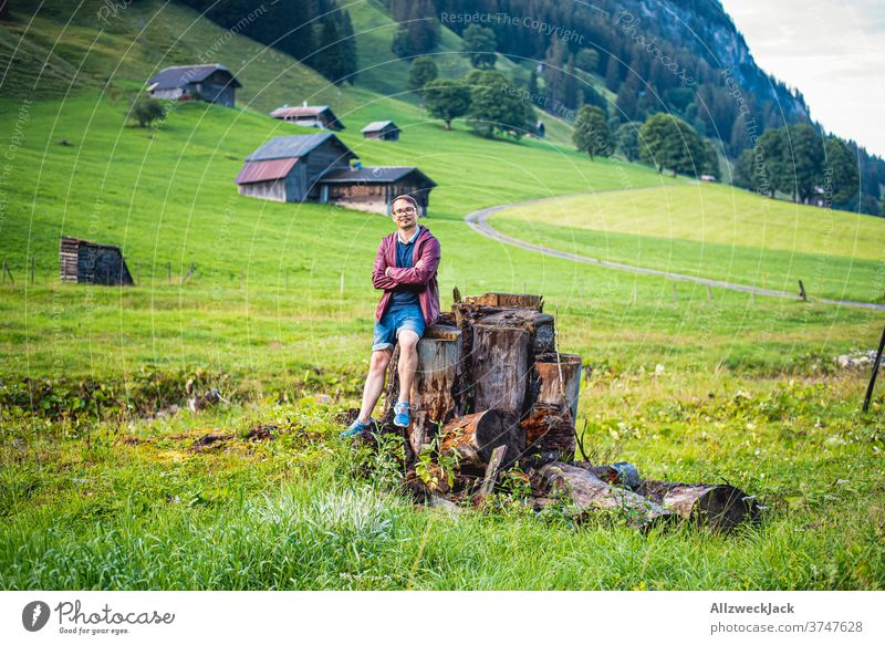 junger Mann mit Brille, Regenjacke und kurzen Hosen sitzt auf einem Baumstumpf auf einer schweizer Alm und schaut in die Kamera Junger Mann Schweiz rasten