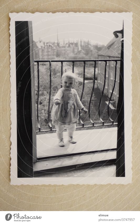 Erinnerungen an die 1960er Jahre - ein schwarz-weißer Fotoabzug mit Büttenrand liegt auf beigefarbenen Papier und zeigt ein kleines Mädchen von einem Balkon aus in das Zimmer guckend