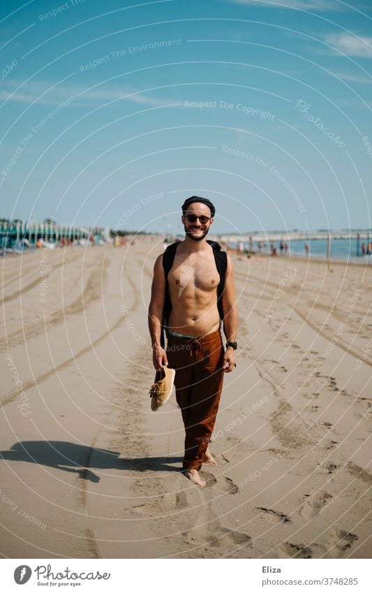 Ein Mann mit freiem Oberkörper steht am Strand und grinst freudig in die Kamera freier Oberkörper Freude Urlaub Tourist Erwachsene Meer nackter oberkörper