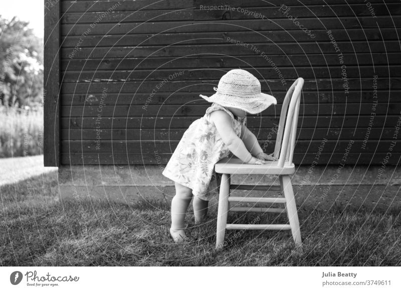 Kleinkind, das einen Sonnenhut trägt und sich an einem Stuhl festhält, um vor einer Scheune zu stehen Baby Kind 0-12 Monate Kindheit klein niedlich entspannt