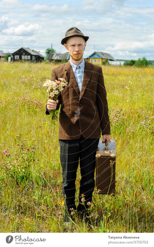 Mann-Hipster mit altem Retro-Koffer und Wildblumen-Bouquet Landschaft cottagecore altehrwürdig Sommer braun Feld grün Jacke retro rustikal reisen jung männlich