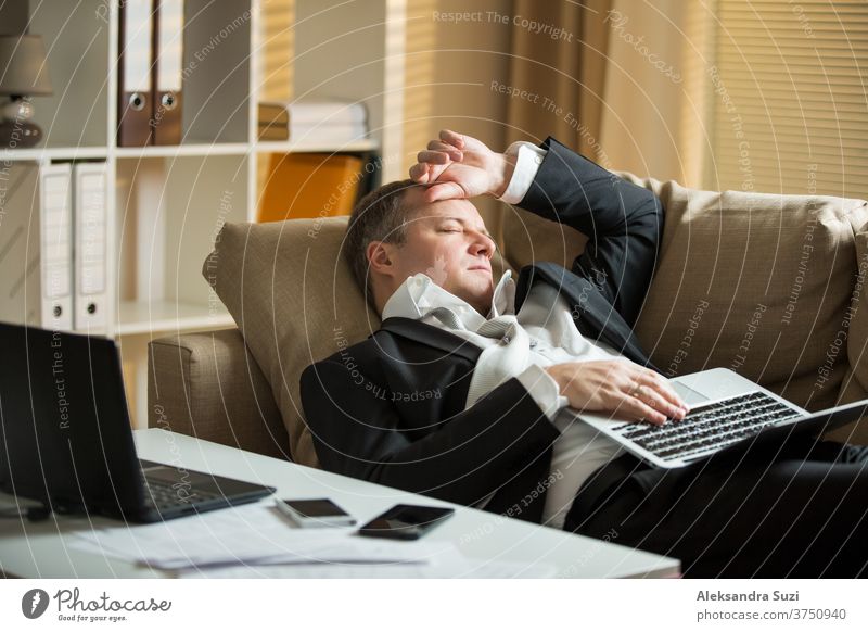 Geschäftsmann im Anzug, auf einer Couch liegend, mit zwei Mobiltelefonen und Laptops, schlafend. Erschöpfter Mann entspannt sich am frühen Morgen im Büro. Verantwortlich arbeitender leitender Angestellter schlief ein.