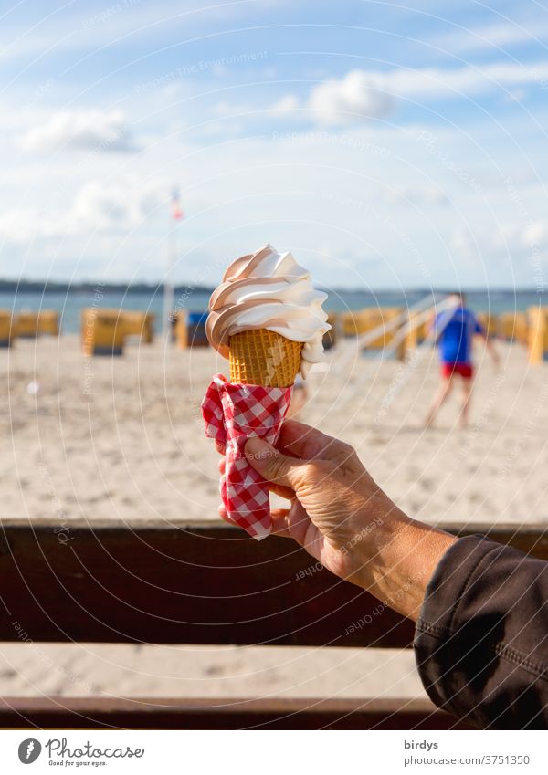 Eine Hand hält eine Eiswaffel mit Softeis.Unscharfer Hintergrund mit Strand , Strandkörben , Meer und einer Person.Schönes Wetter mit Sonnenschein . Blauer Himmel , Wolken