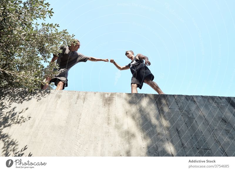 Männliche Freunde auf hoher Mauer in der Stadt stoßen Faust Gruß Männer Zusammensein Freundschaft Le Parkour Training vorbereiten Stein Gebäude Großstadt Wand