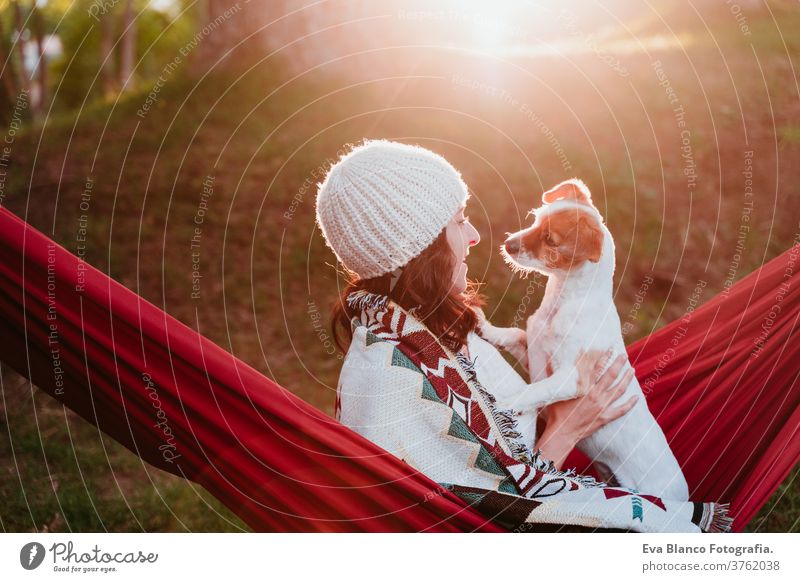 junge Frau entspannt sich mit ihrem Hund in orangefarbener Hängematte. Mit Decke zugedeckt. Zelten im Freien. Herbstsaison bei Sonnenuntergang