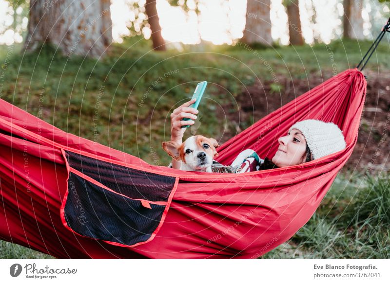 junge Frau entspannt sich mit ihrem Hund in orangefarbener Hängematte. Mit Decke zugedeckt. Benutzung eines Mobiltelefons. Zelten im Freien. Herbstsaison bei Sonnenuntergang