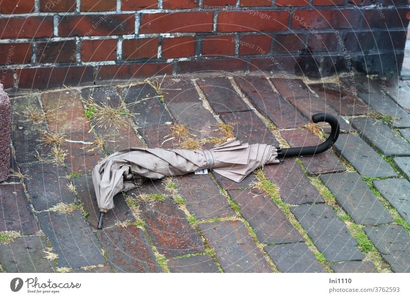 unbrauchbarer, weggeworfener Regenschirm zusammengeklappt verbogen entsorgt beige krumm Pflastersteine vergessen Müll achtlos Gras Wangerooge Ortsmitte