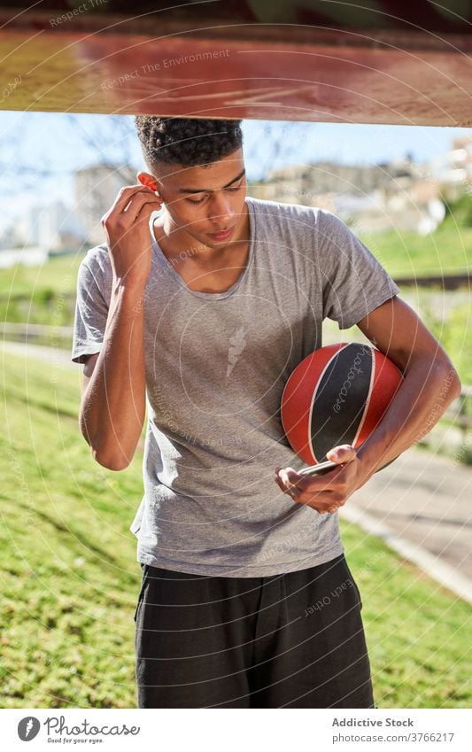 Streetball-Spieler mit Ball beim Musikhören Sportler Smartphone Ohrstöpsel benutzend zuhören Training Pause angezogen ausrichten Athlet jung Mann Basketball