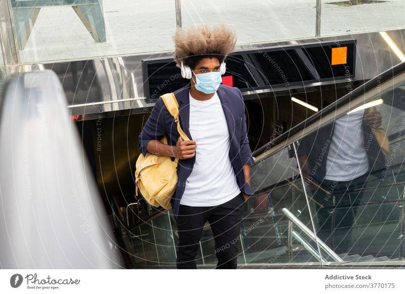 Afrikanischer Mann mit Maske auf einer Rolltreppe mit einer Tasche, während Liste Seuche Coronavirus Pandemie schützend Sicherheit Gesundheit Tourist