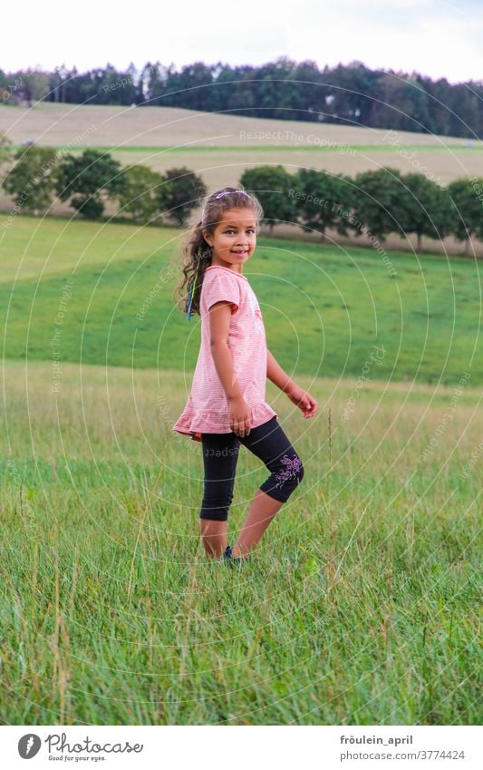 Mädchen auf dem Land Kind lange Haare Wiese grün Bäume Natur Umwelt lächeln Fröhlichkeit fröhlich rosa schwarz Feld Sommer Kindheit Freude Außenaufnahme Gras