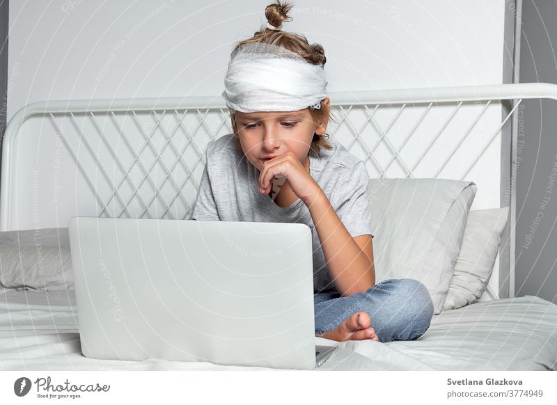 Der kleine kaukasische blonde Junge mit einer Kopfverletzung und einem Verband sitzt auf dem Bett und benutzt einen Laptop. Erholung nach dem Vorfall. Fernkommunikation, Fernunterricht, Technologien und Gadgets im Leben