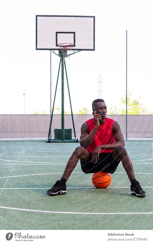 Ethnischer Basketballspieler, der auf dem Spielfeld mit seinem Smartphone spricht Sportler reden Spielplatz Sportpark benutzend Ball sich[Akk] entspannen Sommer
