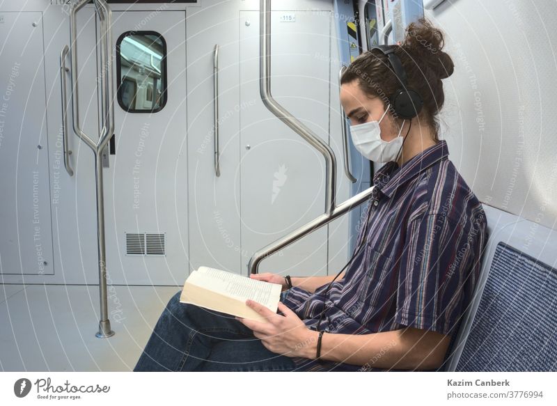 Konzentriert maskiert tausendjährig ein Buch lesen und in der U-Bahn Musik hören unterirdisch Mundschutz Kopfhörer Sitzen Mann jung Schüler Teenager Universität