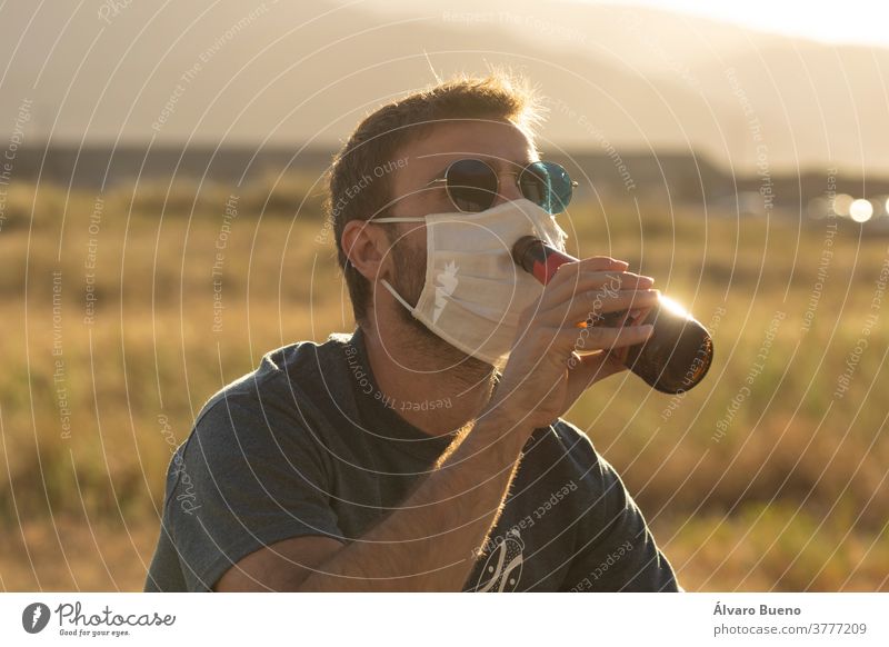 Junger Mann versucht, mit einer schützenden Gesichtsmaske Bier zu trinken. Tag Sonnenuntergang Tageslicht Person jung Erwachsener Sonnenbrille im Freien