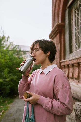 Junge erwachsene Frau mit kurzen Haaren trinkt aus Metall nachhaltiges Wasser im Freien, selektiver Fokus Flasche Hände Nahaufnahme Finger Beteiligung Mädchen