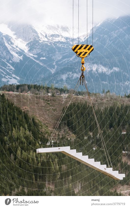 Schwebende Karrieretreppe aus Beton am Kranhaken, präsentiert vor verschneiter Bergwelt Gehänge Kettengehänge hängen Treppe Fertigteil betonelement Baustelle