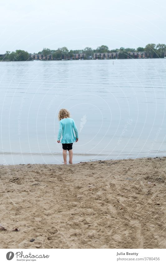 Kleines Mädchen mit lockigem blonden Haar allein am Strand am See Seeufer Sand Wasser Rippeln Kind krause Haare blondes Haar Isolation watend blau weich