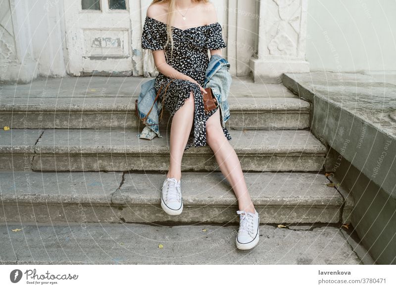 Gesichtslose Aufnahme einer jungen schlanken Frau, die auf einer Treppe im Freien sitzt und eine Kamera in den Händen hält lang Beine Tag Großstadt Hipster Mode