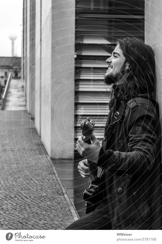 Ein junger Musiker singt im Regierungsviertel Berlin und zupft auf seiner Gitarre Bassgitarre Lied Straßenmusik nachdenklich traurig melancholisch Haare Gebäude