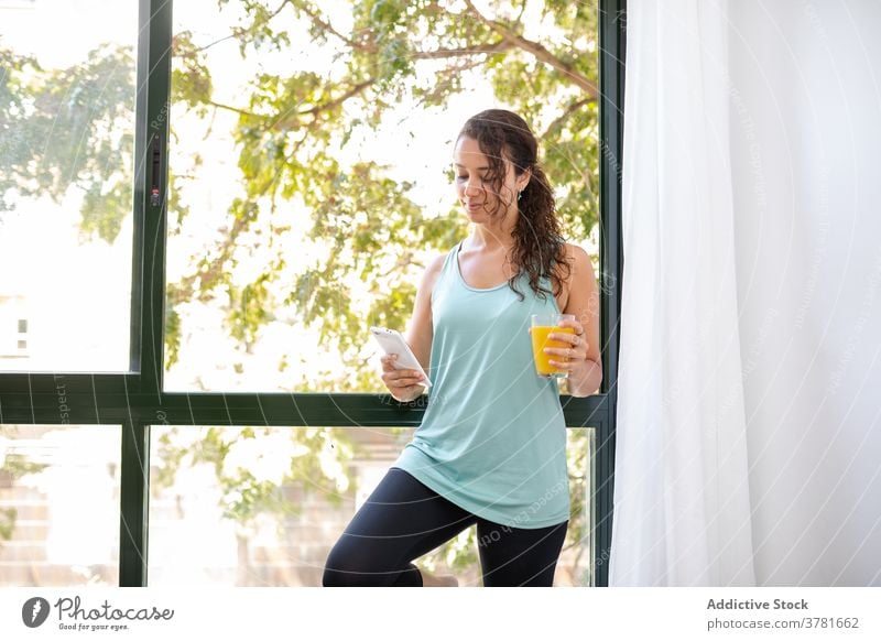 Ruhige Frau mit Saft mit Smartphone zu Hause Morgen orange benutzend genießen Talkrunde soziale Netzwerke Wochenende frisch Glas Fenster heimisch trinken