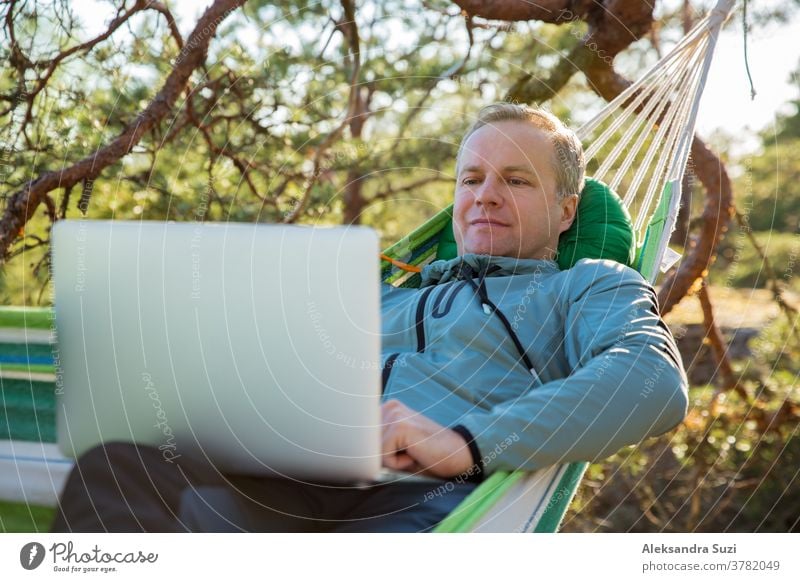 Ein Mann arbeitet an einem Laptop, während er in einer Hängematte im Wald liegt. Selbstisolierung, Freiberuflichkeit, Fernarbeit und Distanzierung. Draufsicht auf die skandinavische Landschaft